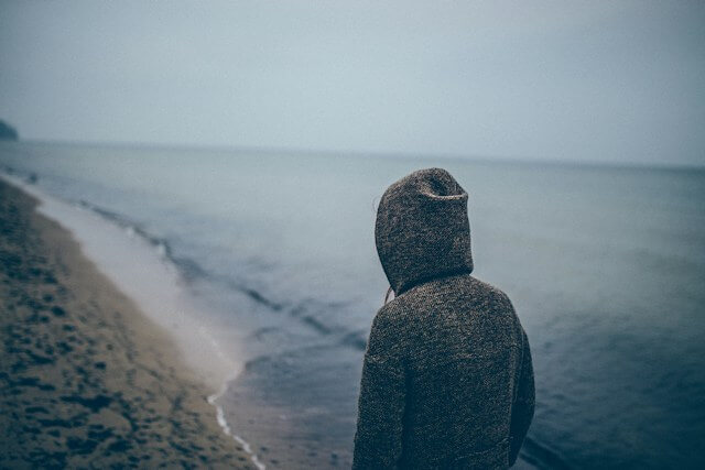 A hooded person standing alone beside a quiet beach