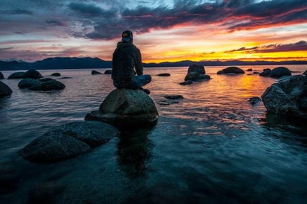 A man with a baseball cap sitting on a rock at the edge of a beach