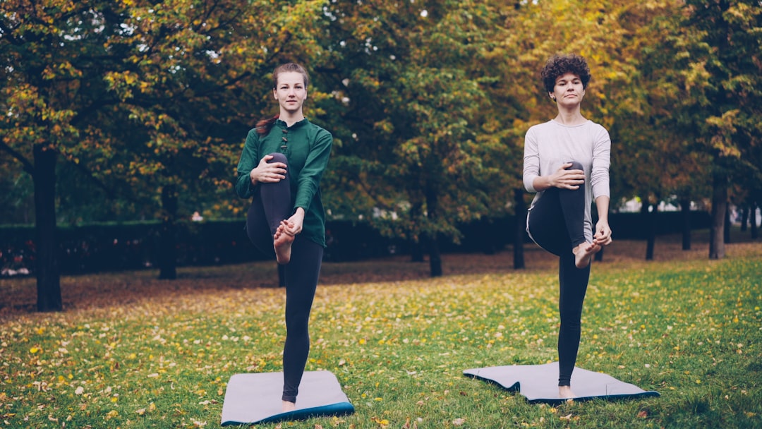 Two people doing gentle stretches on grass in a park with autumn leaves scattered around