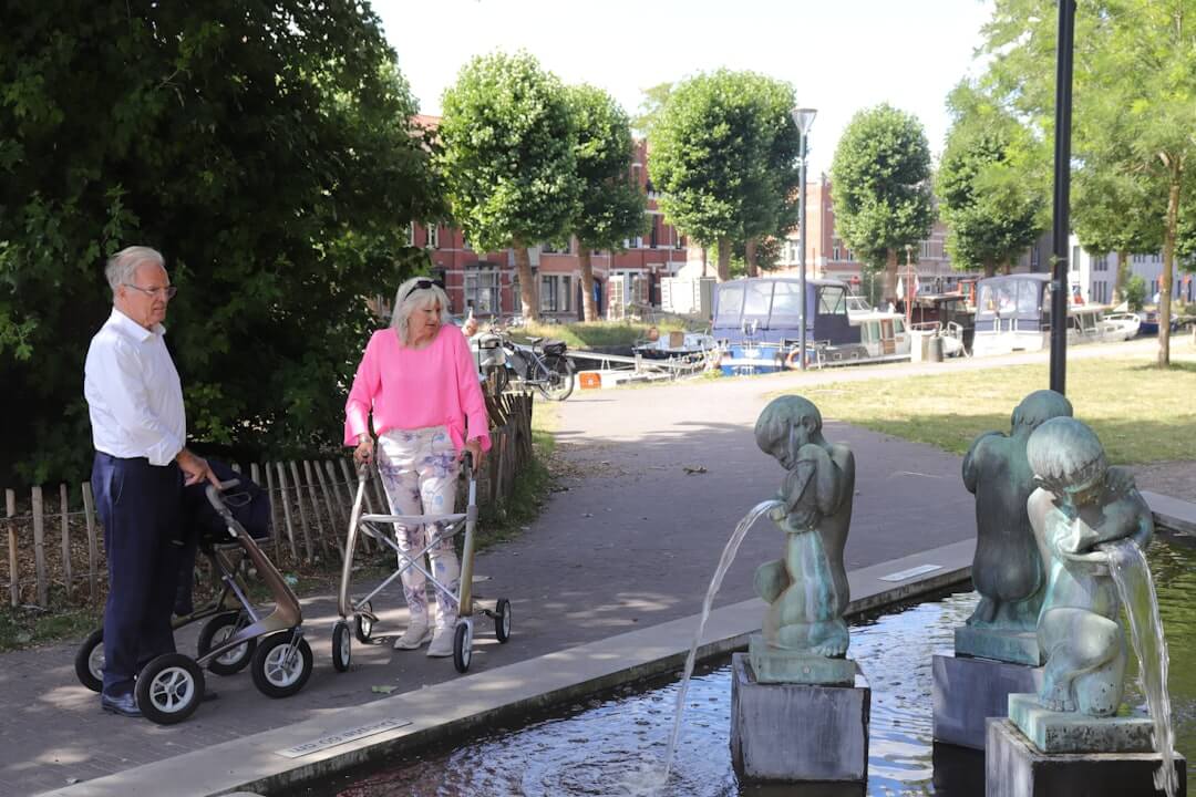 Two grey-haired adults with walking aids beside a fountain on a park path