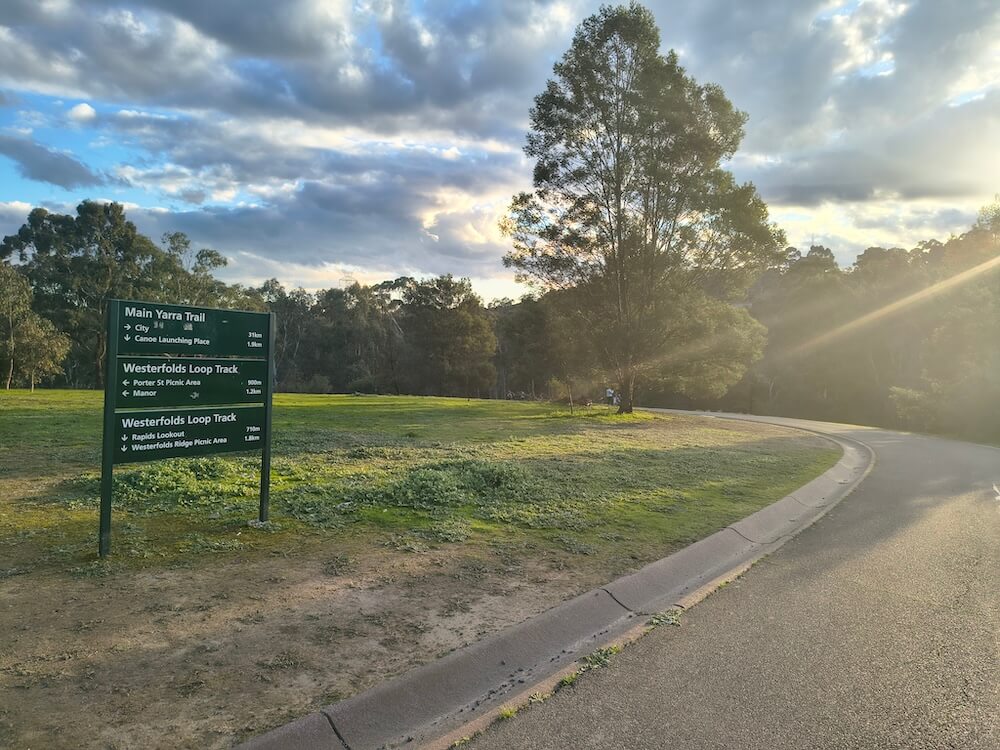 A Main Yarra Trail direction sign beside a sealed path in Westerfolds Park at sunset