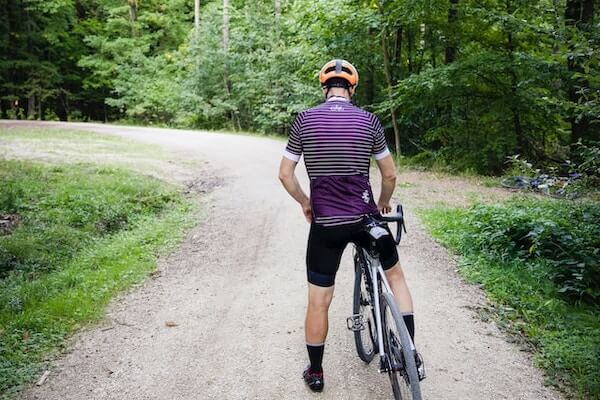 Cyclist wearing a helmet and walking a road bike along a gravel forest track