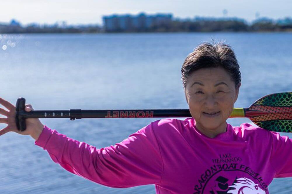 An older dragon boat paddler smiling beside the water with a paddle across her shoulders